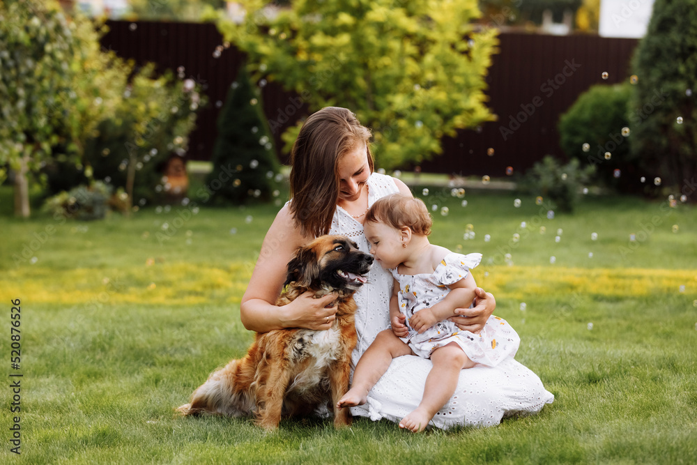 Fototapeta premium Family day, mother's day. Beautiful smiling young mom and baby daughter cuddling happy domestic dog on the backyard lawn.Mother with child girl are having fun with pet outdoors on summer holiday.