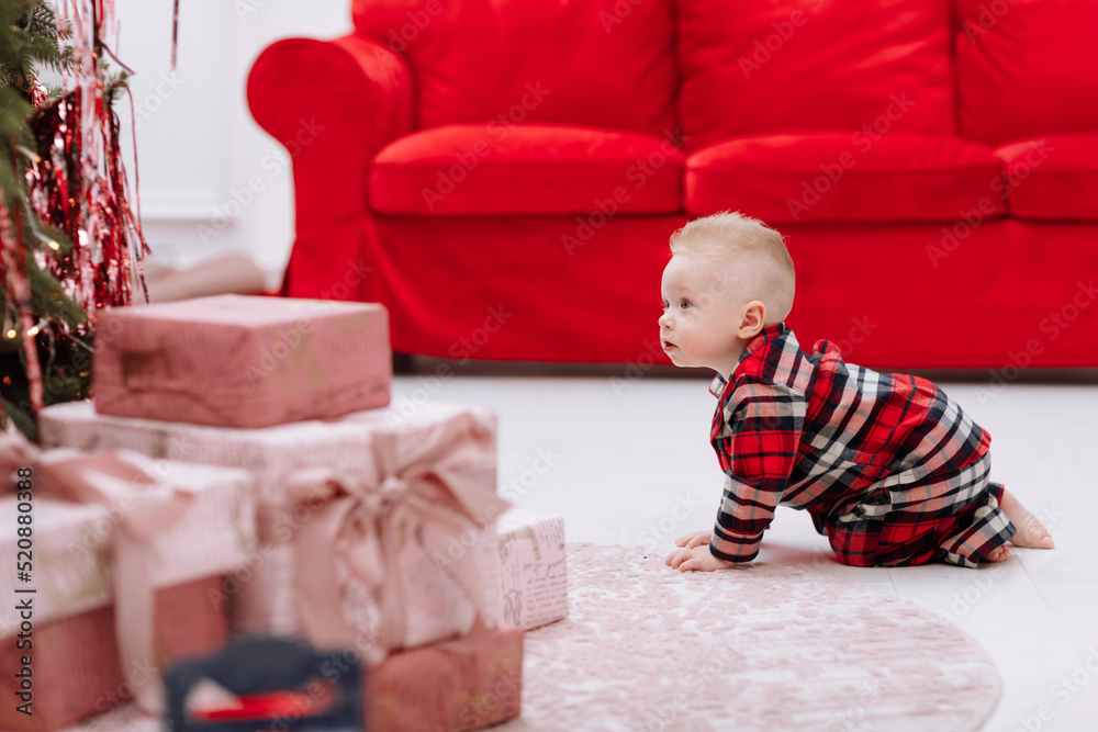 Adorable caucasian baby boy is crawling by the Christmas tree, many festive gift boxed and red sofa at home, cute infant in stylish pyjama. Xmas and New Year family celebrations concept. Adorable caucasian baby boy is crawling by the Christmas tree, many festive gift boxed and red sofa at home, cute infant in stylish pyjama. Xmas and New Year family celebrations concept.