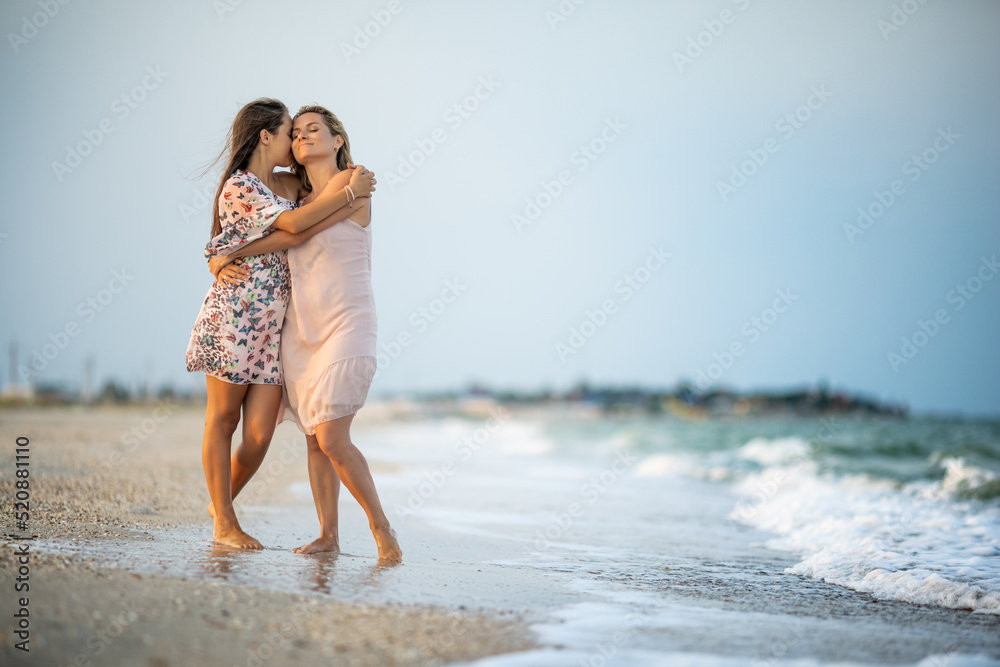 Fototapeta premium A girl in a dress hugs her mom in a dress while walking along the beach near the sea with waves