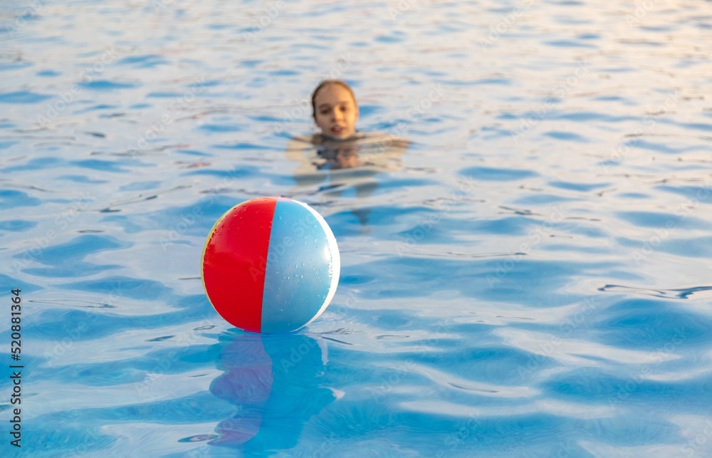 A girl in a bright bathing suit swims with an inflatable ball in a pool with clear water on a summer evening
