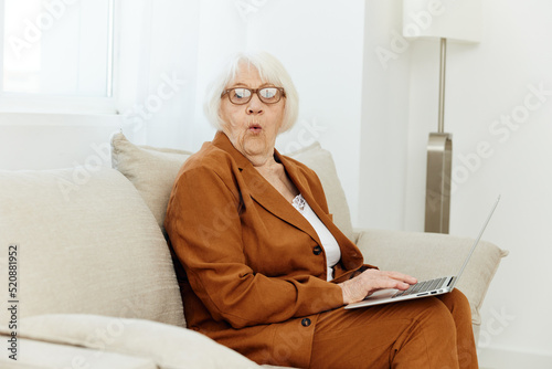 struck with surprise and shock, an emotional woman with age-gray hair sits on the couch with a laptop while working from home and looks at the camera in shock and bewilderment