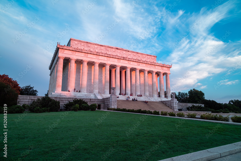 Pink light from sunrise illuminates the east-facing columns and exterior of the Lincoln Memorial in Washington, DC. Several sightseers sit on the marble steps.