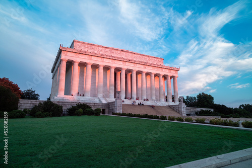 Pink light from sunrise illuminates the east-facing columns and exterior of the Lincoln Memorial in Washington, DC. Several sightseers sit on the marble steps.