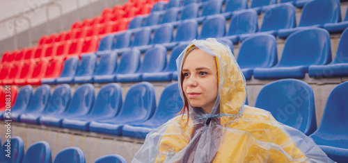 Young woman in raincoat sitting on stadium bleachers alone in rainy weather. Female spectator in wet waterproof coat at street stadium during competitions.