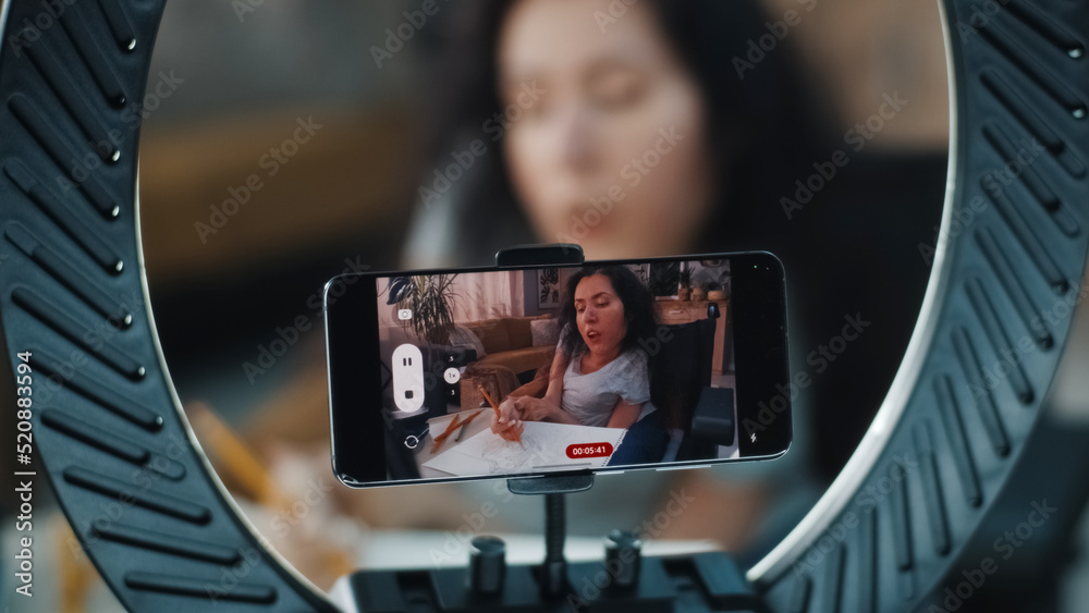Woman in a wheelchair drawing with pencil at the table while filming the process for her blog on phone camera using a tripod, phone holder and ring LED lamp