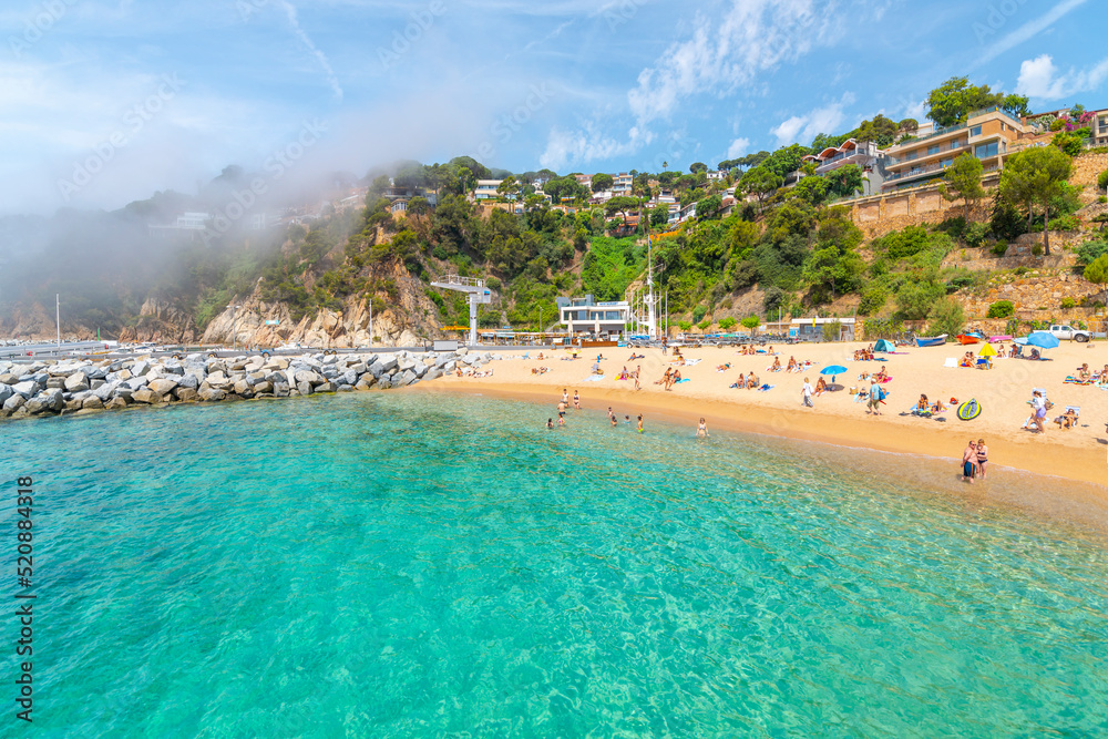 Lloret de Mar, Spain - June 7 2022: View from the clear turquoise sea as the fog breaks at morning along the Cala Santa Cristina sandy beach on the Costa Brava coast in Lloret de Mar, Spain.