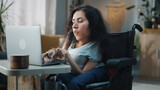 Woman with disability in a wheelchair at home browsing the internet, online shopping or use social media using a laptop at daytime in cozy room