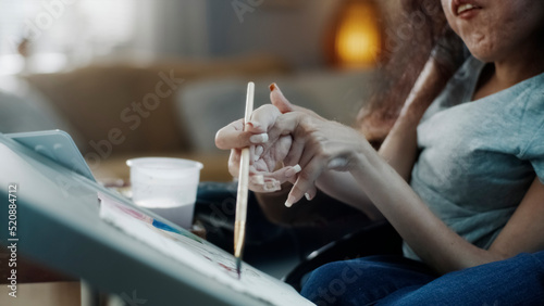 Close up shot of beautiful woman artist in a wheelchair, with spinal muscular atrophy sitting at the table and painting at home in spare time.