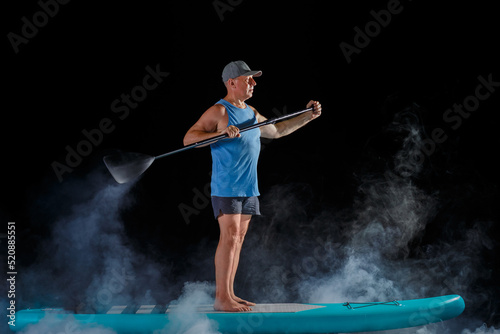 A man in a cap on a sub board with a paddle on a black background in the fog.