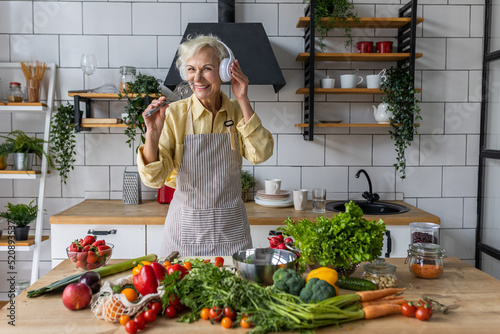 Senior attractive woman listening to music and singing in her headphones in the kitchen. Active lifestyle of mature people, enjoying life, having fun. Using whisk as microphone while cooking food