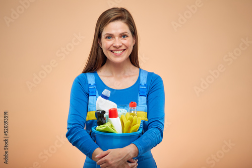 Smiling woman in overalls holding bucket with cleaning products. isolated on beige background.