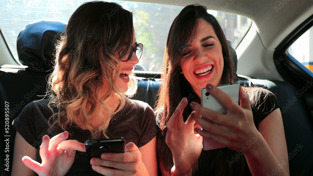 Female friends browsing on their smartphone while riding cab laughing. Women laughing while using cellphone in the back seat of a car