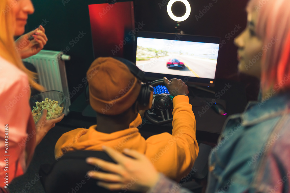 Two teenage girls talking watching teenage boy playing racing game using steering wheel. Gaming setup. Red lighting. . High quality photo