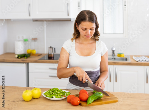 Positive woman is cutting vegetables for salad in the kitchen at home