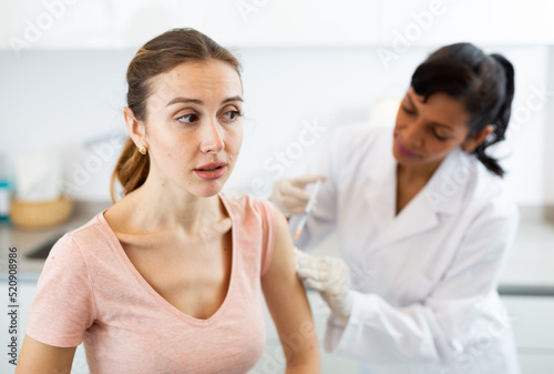 Woman doctor in sterile gloves giving shot to female patient while she sitting on bed with unhappy grimace on face