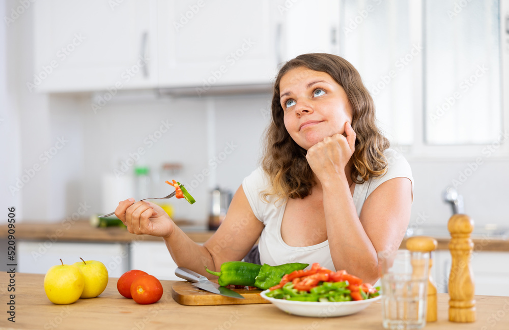 Young positive woman tasting bell pepper during cooking at kitchen, looking away