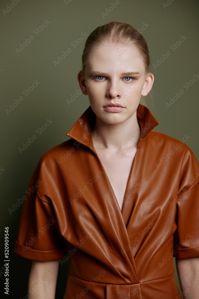 Seasonal Sale offer. Confidence blond young lady in trendy leather trench coat dress looks with blue eyes at camera presenting evening makeup posing isolated on olive green studio wall background Seasonal Sale offer. Confidence blond young lady in trendy leather trench coat dress looks with blue eyes at camera presenting evening makeup posing isolated on olive green studio wall background