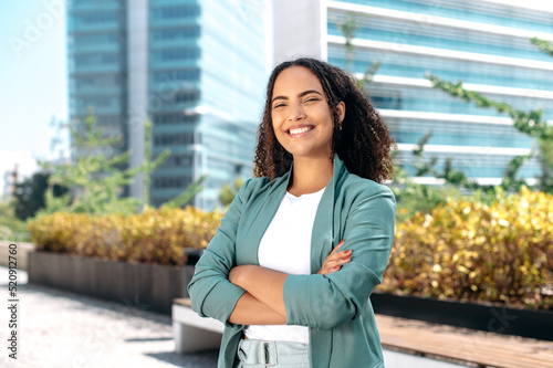 Successful elegant confident young mixed race woman with curly hair, formally dressed, stands outdoors against the background of the business center, with arms crossed, looks at camera, smile friendly