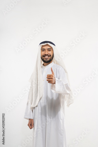an arabian young man in a turban smiling while standing looking at the camera with thumbs up while talking against a plain background photo