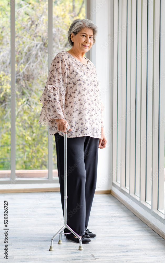 Full body shot Asian old senior happy healthy gray hair female retired pensioner in casual wear standing smiling in hospital hallway with glass windows using support assist walking cane stick walker