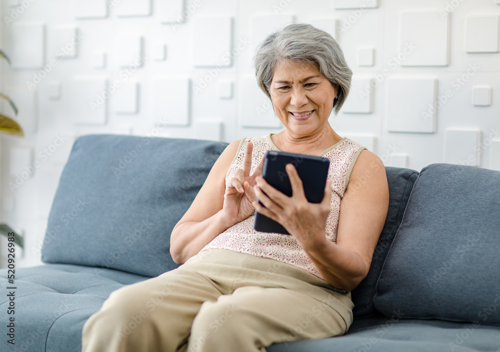 Asian cheerful old senior healthy gray hair female pensioner grandmother sitting smiling on cozy sofa in living room holding using touchscreen tablet computer surfing browsing application online Asian cheerful old senior healthy gray hair female pensioner grandmother sitting smiling on cozy sofa in living room holding using touchscreen tablet computer surfing browsing application online