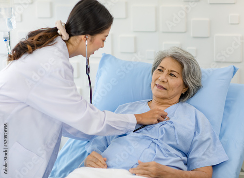 Closeup shot of unrecognizable unknown doctor in white lab coat with stethoscope hand holding comforting supporting old senior unhealthy patient in hospital uniform laying down on bed in ward room
