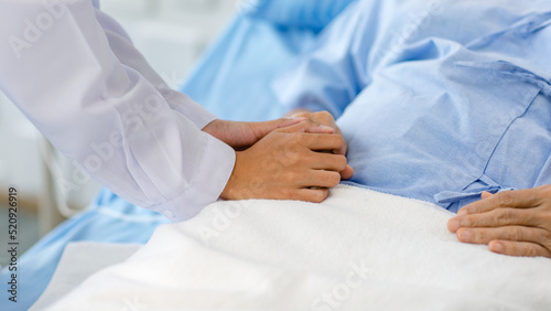 Closeup shot of unrecognizable unknown doctor in white lab coat with stethoscope hand holding comforting supporting old senior unhealthy patient in hospital uniform laying down on bed in ward room