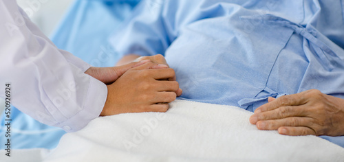 Closeup shot of unrecognizable unknown doctor in white lab coat with stethoscope hand holding comforting supporting old senior unhealthy patient in hospital uniform laying down on bed in ward room