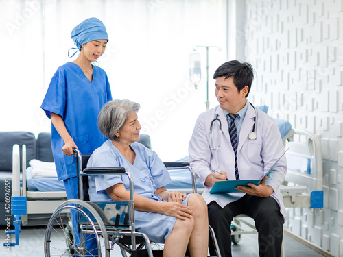 Asian male professional doctor practitioner in white lab coat and stethoscope writing notes when visiting monitoring examining old senior female pensioner patient sitting on wheelchair in hospital