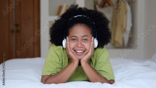 A cute black woman with an afro hairstyle lies on the bed and smiles a snow-white smile. Afro Asian woman listens to music with wireless headphones. The life of a young multiracial woman.