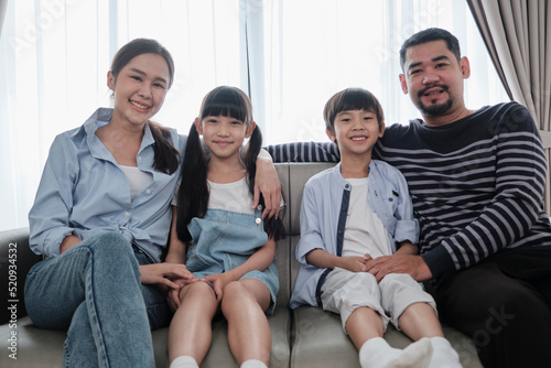 Portrait of Asian Thai family, adult dad, mum with little children happiness home living together, looking at camera, leisure on sofa in white room, lovely weekend and wellbeing domestic lifestyle.
