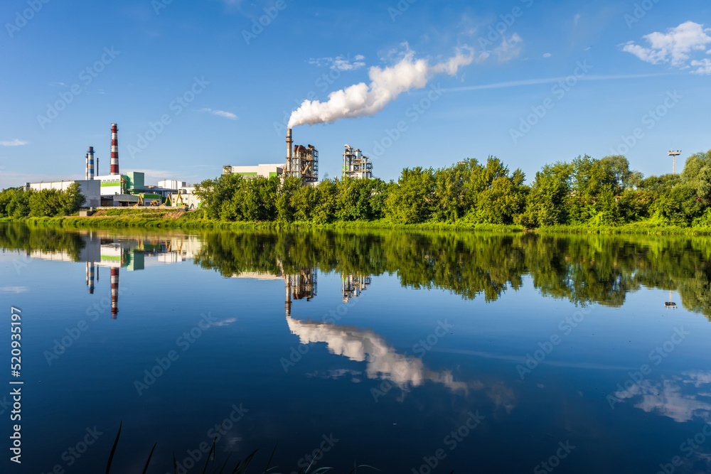 pipes of woodworking enterprise plant sawmill with beautiful reflection in blue water of river. Air pollution concept. Industrial landscape environmental pollution waste of thermal power plant