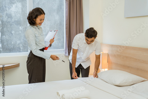 Housekeeping manager carrying clipboard notes while supervising maid cleaning bed in hotel room
