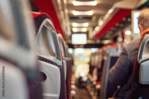 Row of seats in a tourist bus and male passenger out of focus. Selective focus. Travel and commute concept. The interior has bald red color scheme, plastic seats and TV screen. © mark_gusev
