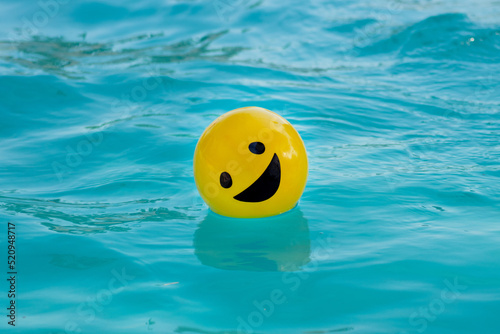 Pelota amarilla con una cara sonriendo flotando sobre el agua azul photo