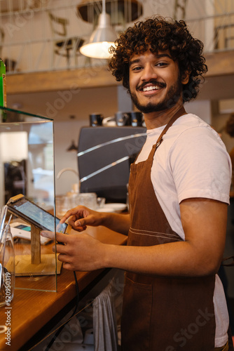 Young indian smiling handsome curly barista working with tablet
