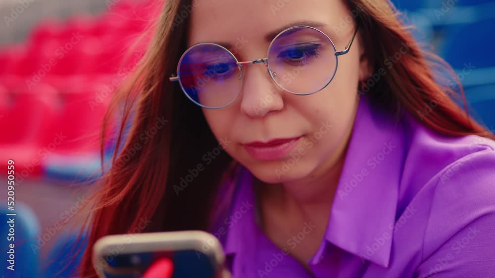 Concentrated adult female with long brown hair in blouse and eyeglasses sitting on seat of blue tribune and messaging on smartphone Concentrated adult female with long brown hair in blouse and eyeglasses sitting on seat of blue tribune and messaging on smartphone