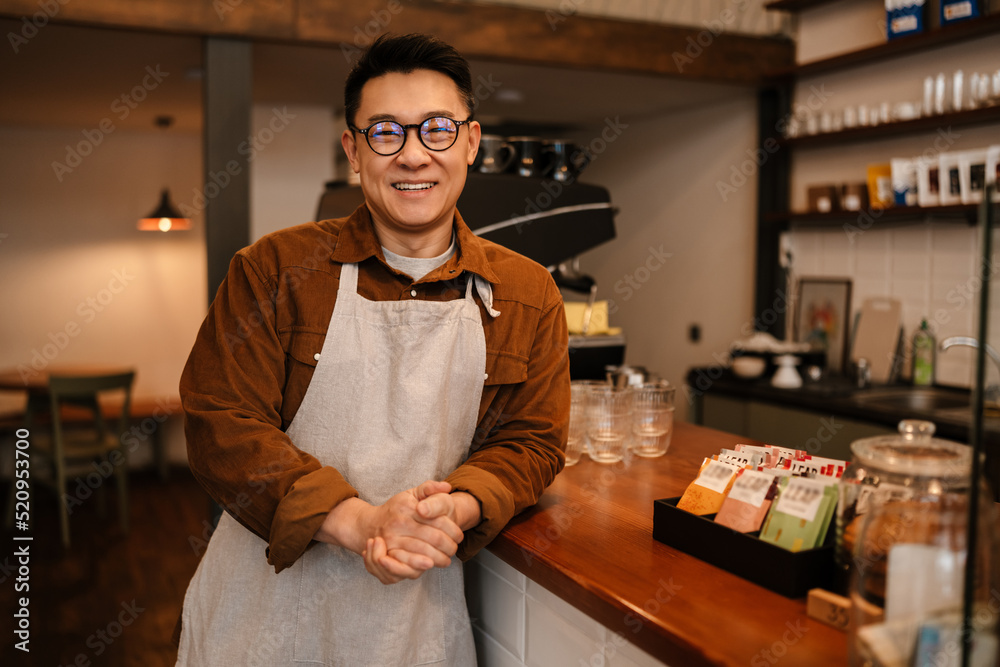 Fototapeta premium Adult asian man wearing apron smiling while working in cafe indoors