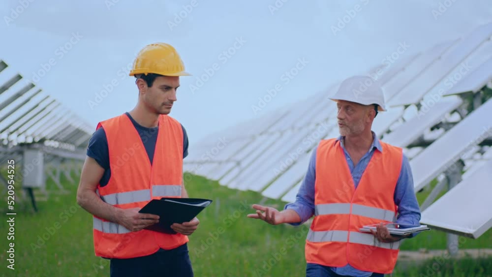 Walking in front of the camera the two charismatic men workers examining solar batteries and communicating of business they walking between rows Walking in front of the camera the two charismatic men workers examining solar batteries and communicating of business they walking between rows