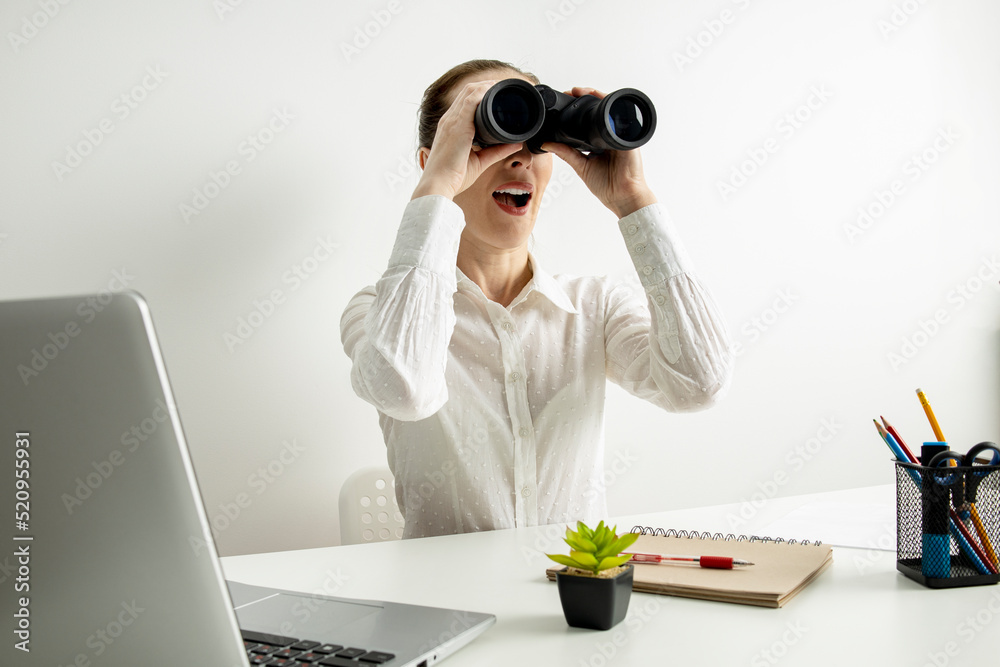 Surprised young woman looking through binoculars while sitting at office desk Surprised young woman looking through binoculars while sitting at office desk