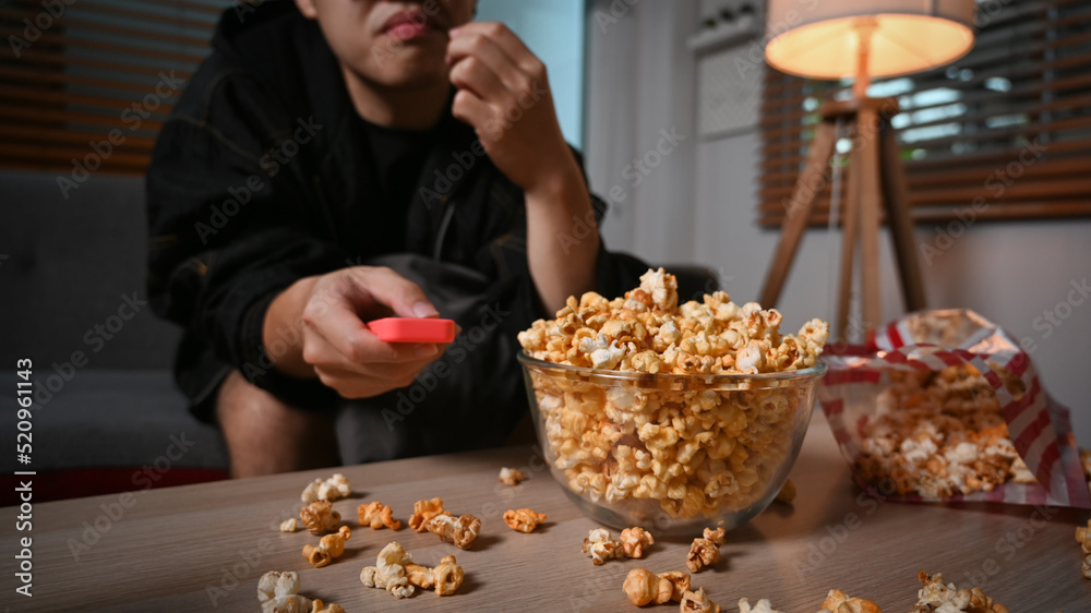 Joyful young asian man eating popcorn and watching TV at home in cozy living room