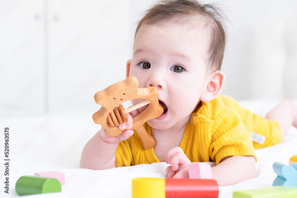 healthy asian baby girl in yellow bodysuit plays with wooden toy teether for teeth on white bedding healthy asian baby girl in yellow bodysuit plays with wooden toy teether for teeth on white bedding