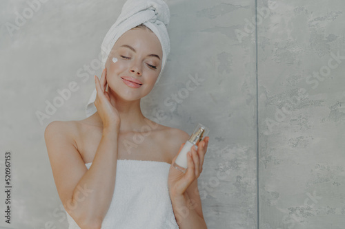 Pretty young woman holds bottle of body lotion  applies face cream on complexion  stands with closed eyes  wears bath towel on head  poses against grey wall. Getting pleasure from skin pampering