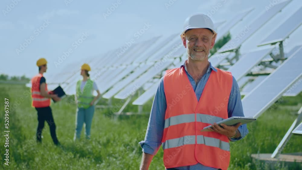 Good looking man engineer holding digital tablet in hands in front of the camera and wearing safety equipment and helmet he posing in the middle of photovoltaic solar farm Good looking man engineer holding digital tablet in hands in front of the camera and wearing safety equipment and helmet he posing in the middle of photovoltaic solar farm