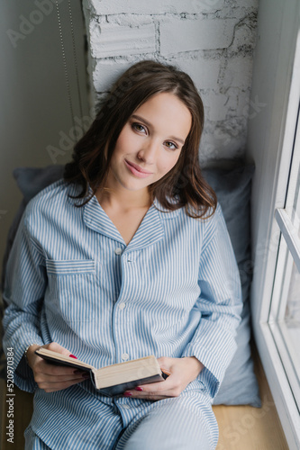 Beautiful relaxed woman with carefree expression, dressed in nightclothes, reads book on window sill in morning, enjoys spare time, likes her hobby. Vertical shot of teenage girl with literature photo