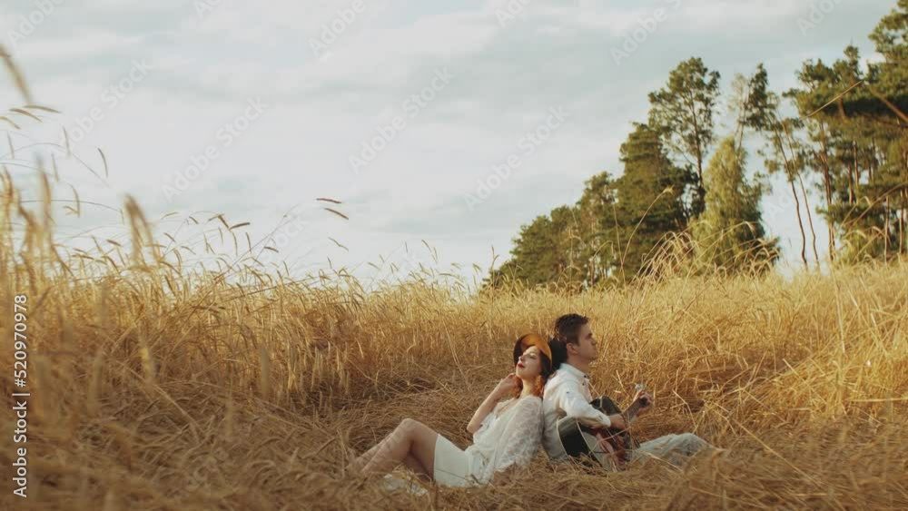 Attractive couple in the field of spikelets slow motion. Woman leans against the boy's back. Guy playing guitar girl listening. Attractive couple in the field of spikelets slow motion. Woman leans against the boy's back. Guy playing guitar girl listening.