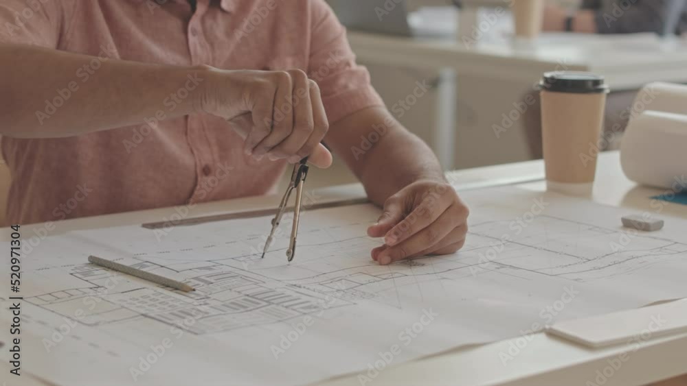 Cropped slowmo of Biracial male architect using compass while drawing plan of building on rolled out sheet of paper in office