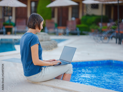 Young business woman working at computer by hotel pool. Young lady downshifter working at laptop and enjoys and relaxed environment, working day. Online freelance work on vacation.