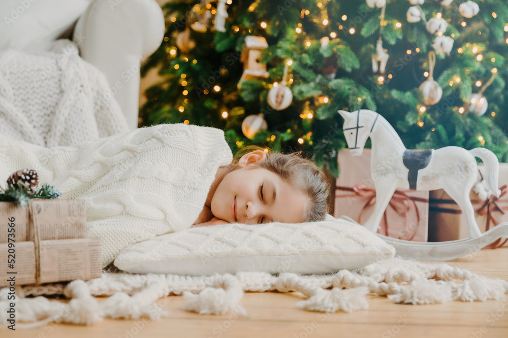 Charming girl sleeps on soft white pillow on floor against decorated New Year tree, has pleasant dreams, surrounded with toy horse and gift boxes. Children, rest and winter holidays concept. Charming girl sleeps on soft white pillow on floor against decorated New Year tree, has pleasant dreams, surrounded with toy horse and gift boxes. Children, rest and winter holidays concept.