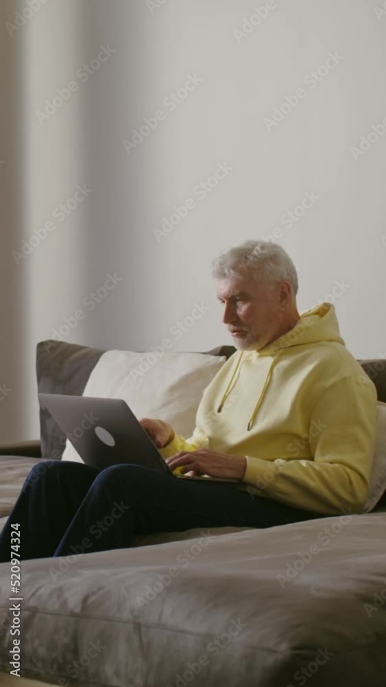 An elderly man smiling starts video call, waving hello sitting on a sofa at home An elderly man smiling starts video call, waving hello sitting on a sofa at home